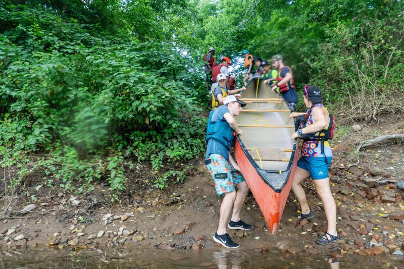 A group of people are carrying a red canoe up a muddy bank from the water. The canoe is being held by two people, one on each side, while others are helping from behind. The bank is covered in rocks and dirt, and there are trees and bushes in the background.